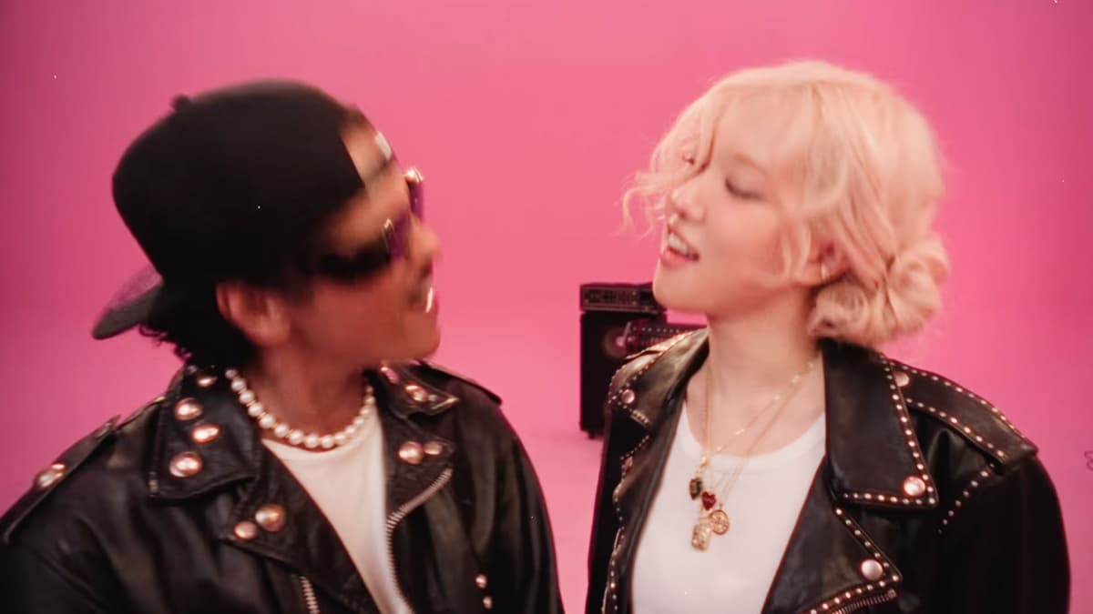 Still from the APT. music video: Bruno Mars in a backwards black cap, dark sunglasses, pearl necklace, and studded black leather jacket on the left, face to face with ROSÉ in a studded leather jacket over a white tee on the right, in a pink studio with a small black amp between them