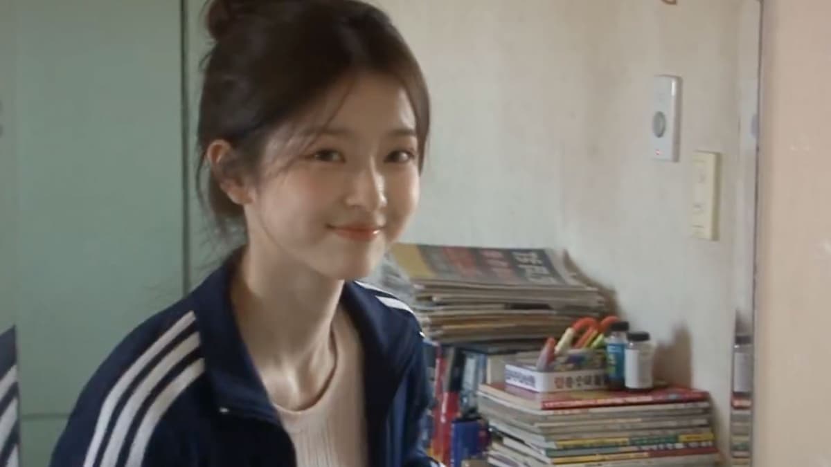 A young woman with her hair in a loose bun, wearing a navy track jacket with white stripes, smiles at the camera in a small bedroom beside stacks of magazines and a pencil cup, in soft, warm, slightly blurred camcorder-style footage
