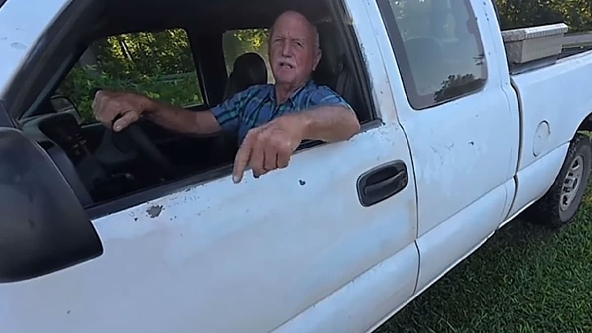 Still from the original TikTok: an older man in a blue plaid shirt leans out the window of a white pickup truck parked on grass by trees, one hand on the wheel and the other pointing at the camera