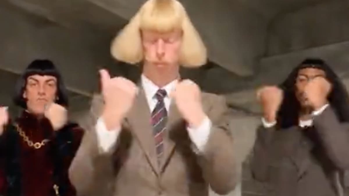 AI-generated still of three deadpan men dancing in sync in a concrete hallway: a blonde man with a blunt Lord Farquaad bob in a tan suit and striped tie in the center, flanked by two men in dark bobs, one in a red velvet tunic with a gold chain
