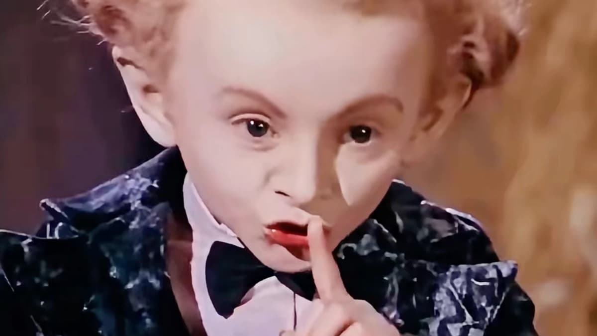 AI-generated close-up of a small man with frizzy ginger hair, a pale face, a black bow tie, a white shirt, and a dark velvet jacket with a pale marbled pattern, holding a finger to his lips, with blurred blonde hair at the right edge of the frame
